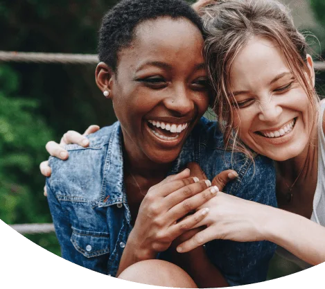 Casal de mulheres abraçadas e sorrindo, uma é negra de cabelos curtos, a outra loira e está usando um coque.