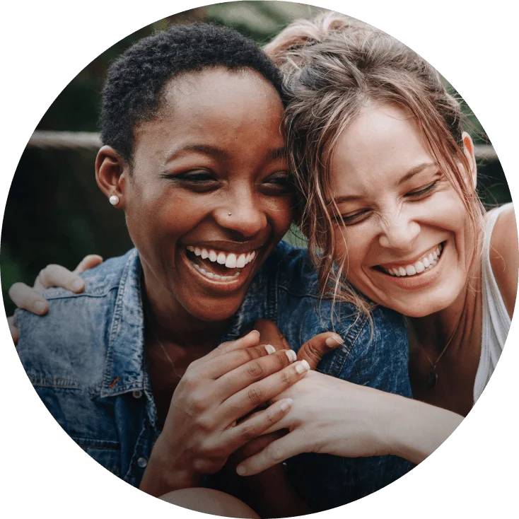 Casal de mulheres abraçadas e sorrindo, uma é negra de cabelos curtos, a outra loira e está usando um coque.